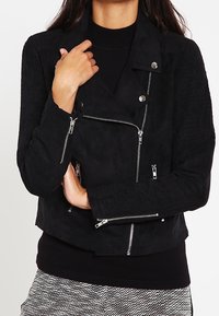 Veste de moto en daim noir avec des détails de zip argentés, un design ajusté et une texture côtelée sur les épaules. Équipée de boutons-pression.