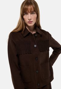 Veste en laine marron avec des franges sur la poitrine, deux poches et fermeture à boutons. Comprend un col et un tissu texturé.