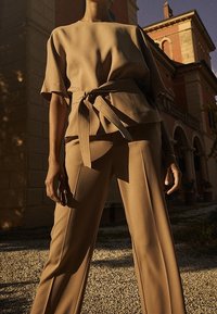 En person klädd i en beige bälte i kortärmad topp och vida byxor står utanför en historisk byggnad i varmt solsken.