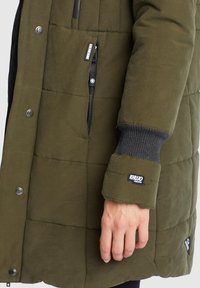 Olivgrön puffjacka med quiltat mönster, utrustad med en dragkedjeficka, ribbade ärmslut och märkespuffar på ärmen.