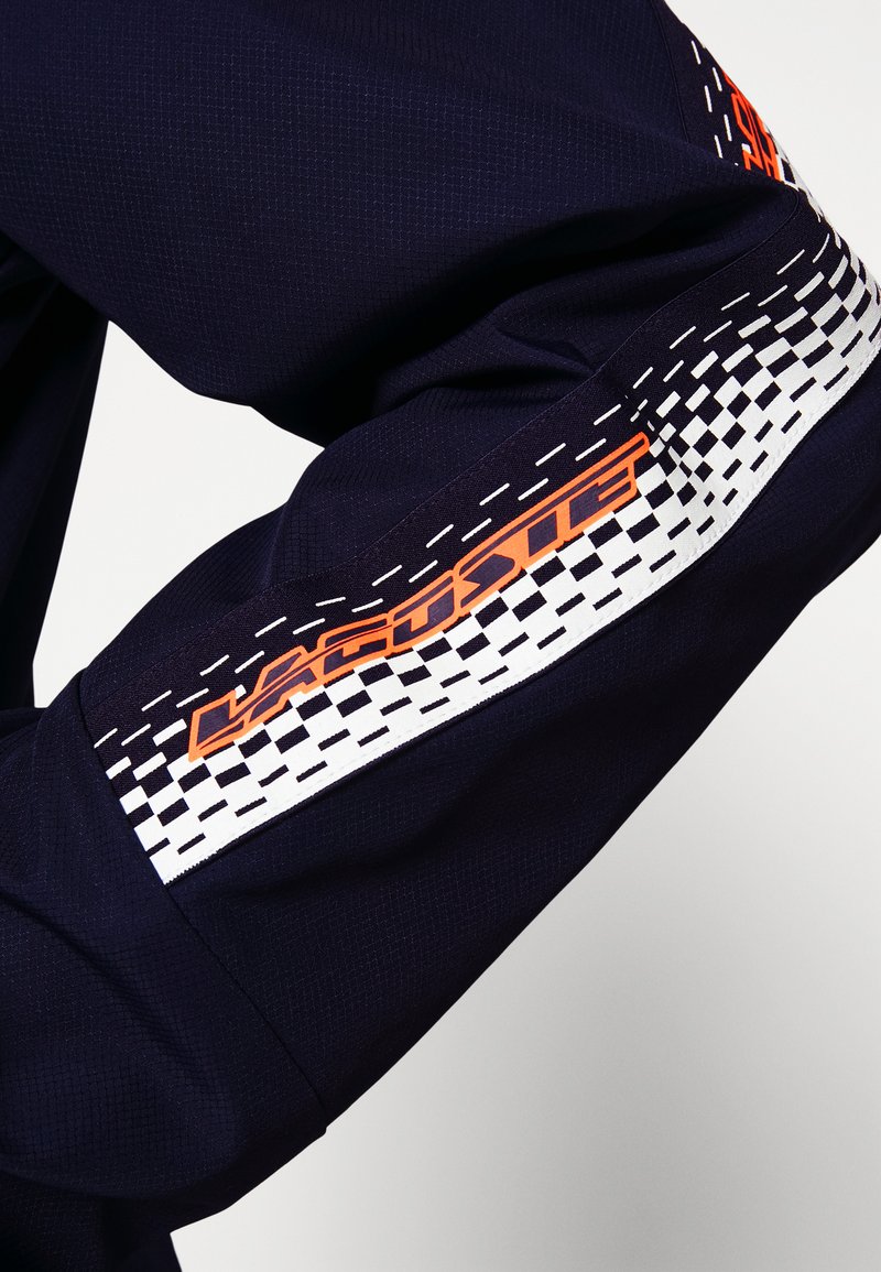 Granatowy rękaw z tkaniny z teksturowanym wzorem, z pomarańczowym logo oraz białym geometrycznym wzorem z czarnymi akcentami w pobliżu mankietu.