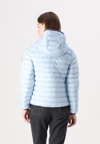 Lichtblauwe pufferjack met een capuchon, voorzien van een gestikte textuur en elastische manchetten. Een contrasterende bies is zichtbaar vlakbij de capuchon.