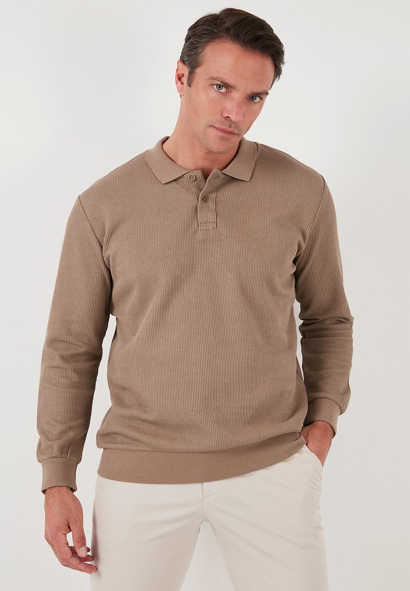 Polo a maniche lunghe in tessuto a coste di colore marrone chiaro con colletto, patta con tre bottoni e polsini aderenti. Abbinato a pantaloni di colore chiaro.