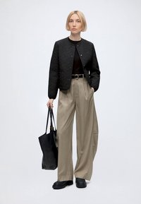 Femme aux cheveux blonds courts porte une veste matelassée noire, un haut noir, un pantalon beige à jambes larges, des chaussures noires et tient un grand sac fourre-tout noir.