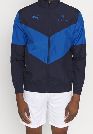 Veste de sport zippée bleu marine et bleue avec logo Puma et texte Italia, portée avec un short blanc par une personne debout.