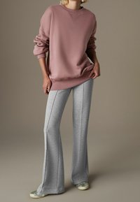 Persoon draagt oversized mauve sweatshirt, lichtgrijze uitlopende broek en zilveren sneakers, staand tegen een effen taupekleurige achtergrond.