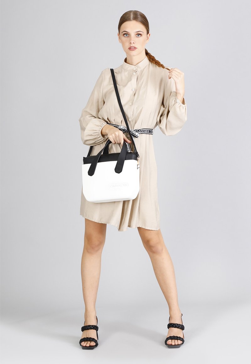 Abito beige con bottoni, maniche voluminose e vita cinte. La modella tiene una borsa bianca con dettagli e cinghie nere. Sandali neri con strappi.
