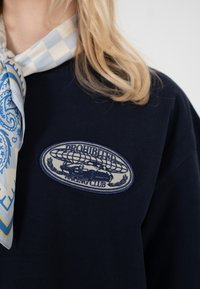 Personne blonde portant un sweat-shirt bleu marine avec un patch ovale montrant une voiture et le texte "Prohibited Racing Club" ainsi qu'une écharpe bleue à carreaux avec un motif cachemire.