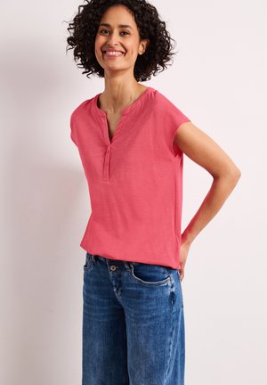 Personne souriante aux cheveux bouclés portant un haut corail à manches courtes boutonné et un jean en denim bleu, debout devant un fond clair.