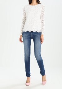 Haut long à manches en dentelle blanche avec ourlet festonné, assorti à un jean bleu ajusté et des talons hauts rose clair. Texture de dentelle douce et délicate.