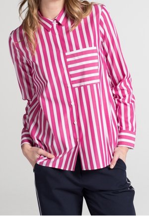 Kvinde iført en lyserød og hvid skjorte med lodrette striber, knapper foran og en matchende stribet brystlomme samt mørkeblå bukser.