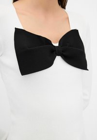 Haut blanc avec un grand nœud en tissu noir décoratif au niveau de l'encolure, confectionné dans un matériau lisse et extensible. Encolure carrée.