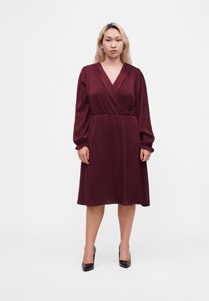 Femme aux cheveux clairs portant une robe bordeaux à manches longues, longueur genou, et des talons hauts foncés, debout devant un fond blanc uni.