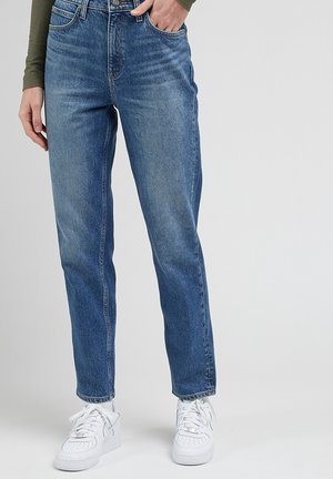 Persoon die blauwe jeans met rechte pijpen en witte sneakers draagt, staat met één hand in de zak tegen een neutrale achtergrond.