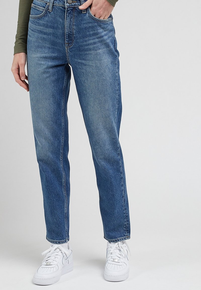 Personne portant un jean droit bleu et des baskets blanches, debout avec une main dans la poche devant un fond neutre.