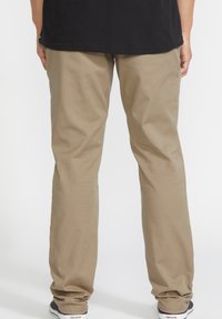Pantalones de algodón beige con un corte relajado, piernas rectas y costuras visibles. Llevados con zapatos negros con suelas blancas y acentos de logo.