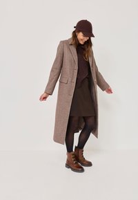 Brązowy długi płaszcz z wełny z kołnierzem, w połączeniu z ciemnym swetrem i spódnicą. Brązowe sznurowane botki dopełniają look.