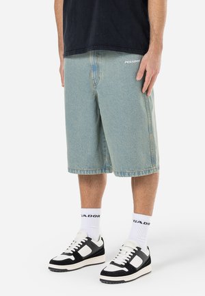 Pantaloni shorts in denim azzurro chiaro, lunghi fino al ginocchio, con cuciture visibili e un logo. Indossati con sneakers nere e calzini bianchi riportanti il nome del marchio.