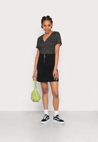 Vestido preto de flores de manga curta com decote em V, apresentando uma saia preta e um detalhe de fecho prata, combinado com ténis pretos e uma bolsa verde.