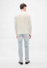 Gaiša krēma džemperis no mīksta, strukturēta auduma ar bezšuvju aizmuguri un ribotiem aproču galiem, kombinēts ar gaiši zilām džinsu biksēm un tumšām kurpēm.