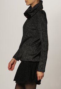 Stickad svart och grå tröja med hög kappa-hals, figurnära silhuett och textilierad tyg. Bärs med en svart kjol med spetsdetaljer.