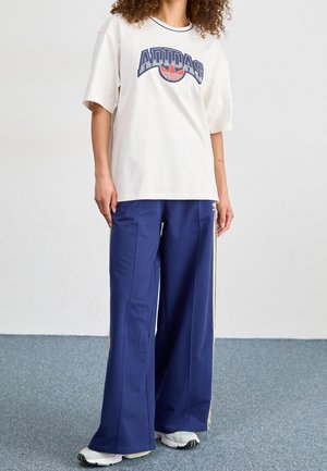 Vit oversized t-shirt med blå Adidas-logotyp, kombinerad med blå vida byxor med gula sidostrippar. Bärs med vita sneakers.