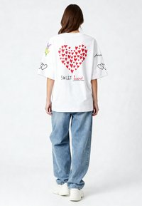 Balta per didelė T-marškinė su raudonų širdžių raštais, tekstu "SWEET heart" ir gėlių grafika. Suderinta su šviesiai mėlynomis plačiomis džinsomis ir baltomis sportbačiais.