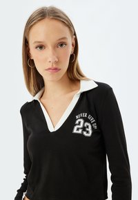 Musta pitkähihaisen paita, jossa on valkoinen kaulus. Paidassa on valkoiset tekstit "ÄLÄ LUOVUTA!" ja "23". Pehmeä kangas, rento istuvuus.