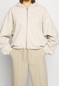 Femme portant un sweat à capuche beige zippé avec poignets élastiques et taille à cordon, associé à un pantalon beige à fines rayures, les mains dans les poches.