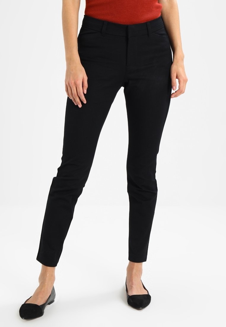 Černé kalhoty na míru se slim fit, s hladkým povrchem a standardními kapsami, v kombinaci s černými plochými botami.