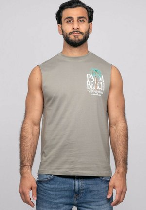 Homme barbu portant un débardeur gris avec le texte "Palm Beach" et un motif de palmier, associé à un jean bleu, sur un fond uni.