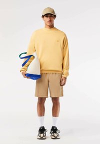 Sudadera amarilla, pantalones cortos beige, gorra beige, calcetines blancos y zapatillas con una suela estampada. Sosteniendo una bolsa con acentos en azul y verde.