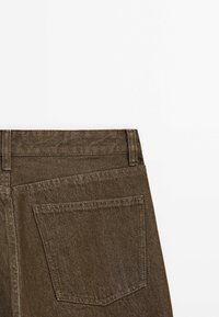 Achteraanzicht van bruine denim broek met tailleband, lussen voor een riem en een enkele achterzak op een witte achtergrond.