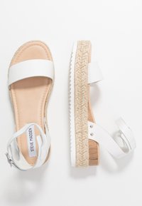 Sandales espadrilles blanches avec une semelle intérieure beige. Elles sont dotées de larges brides, d'une semelle en jute en pente et de détails en métal sur la bride de la cheville.
