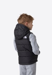 Svart dunvest med hette, med syddede paneler og en liten hvit logo på venstre skulder, brukt over en grå langermet skjorte.