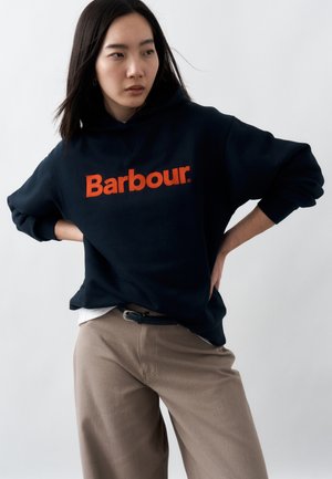 Young woman with long dark hair wearing a navy blue hoodie with orange "Barbour" text, beige pants, standing with hands on hips.