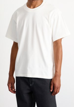 Homme portant un t-shirt blanc uni à manches courtes et un jean noir, debout devant un fond blanc uni.