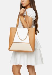 Tote-väska i tan och krämfärg med en strukturerad design, som har en guldkedjeaccent och lädertextur. Handtagen är säkrade för bärande.