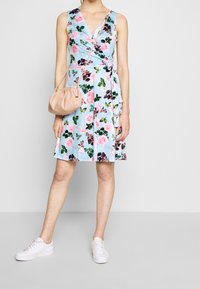 Robe portefeuille florale en bleu clair avec des motifs de roses roses et vertes ; assortie à un sac à main beige et des baskets blanches.