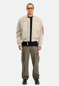 Chaqueta bomber beig con hombros de textura de sherpa, cierre de cremallera y dos bolsillos delanteros. Combinada con una camiseta negra y pantalones cargo oliva.