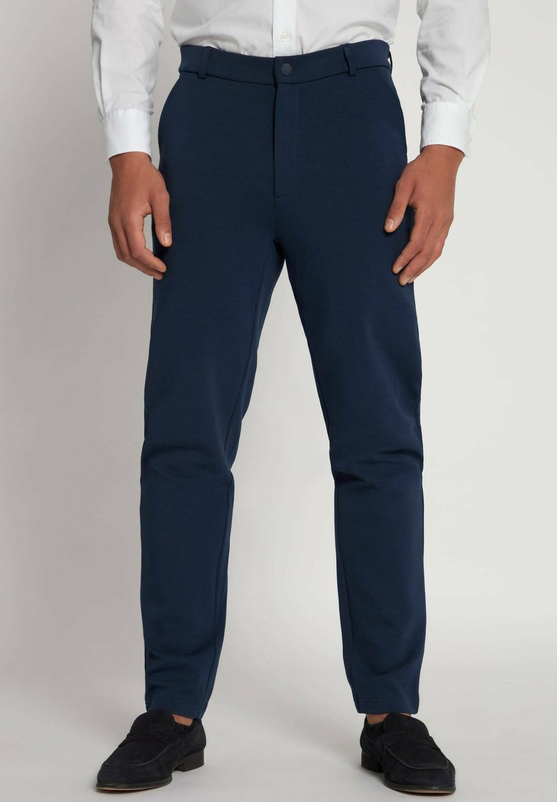 Marineblå bukser lavet af strækbart stof, med en slim fit, bæltesløjfer og lukning med frontknap, parret med sorte sko.