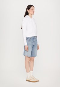 Biely skrátený hoodie s šnúrkami, skombinovaný s svetlomodrými denimovými šortkami a béžovými teniskami s gumovými podrážkami. Model má dlhé vlasy.