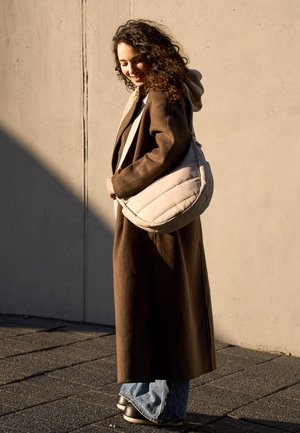 Kvinde med krøllet hår iført en lang brun frakke og blå jeans, bærende på en stor beige skuldertaske, stående foran en betonvæg.