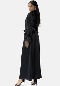 Zwarte maxi-jurk met lange ballonmouwen en een tailleband. Gladde stof met een vloeiende silhouet. Draagt zwarte sandalen met bandjes.