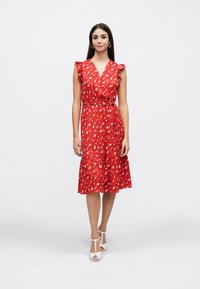 Rochie roșie cu imprimeu floral, cu mâneci cu volane, design tip petrecut și fustă cu volane suprapuse. Are talie ajustată și este asortată cu sandale albe cu bretele.