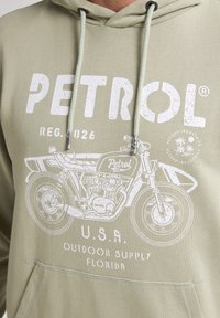 Sudadera verde clara con capucha y cordón. Presenta un gráfico blanco de una motocicleta y el texto "PETROL". Tejido suave con un corte relajado.