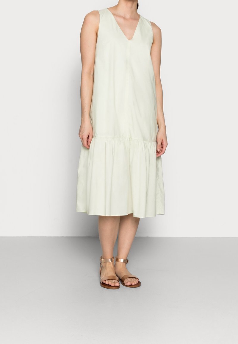 Femme portant une robe midi sans manches couleur crème claire, avec un col en V et un ourlet froncé, associée à des sandales plates à bride cheville couleur beige.