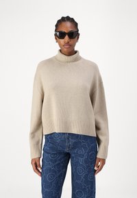 Beiger, gerippter Rollkragenpullover mit überschnittenen Schultern, kombiniert mit dunkelblauen Jeans mit Floral-Muster.