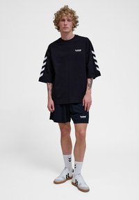Zwarte oversized t-shirt met wit logo, gecombineerd met zwarte korte broek. G gedragen met witte sneakers met zwarte strepen, en witte sokken met zwarte accenten.