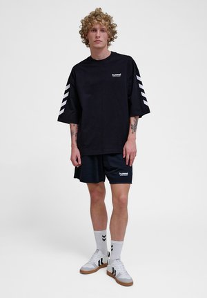 Camiseta negra de gran tamaño con logo blanco, combinada con pantalones cortos negros. Llevada con zapatillas blancas con rayas negras y calcetines blancos con detalles negros.